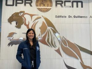 Rachelle Martínez Ramírez frente a un múro que tiene el logo y mascota del Recinto de Ciencias Médicas