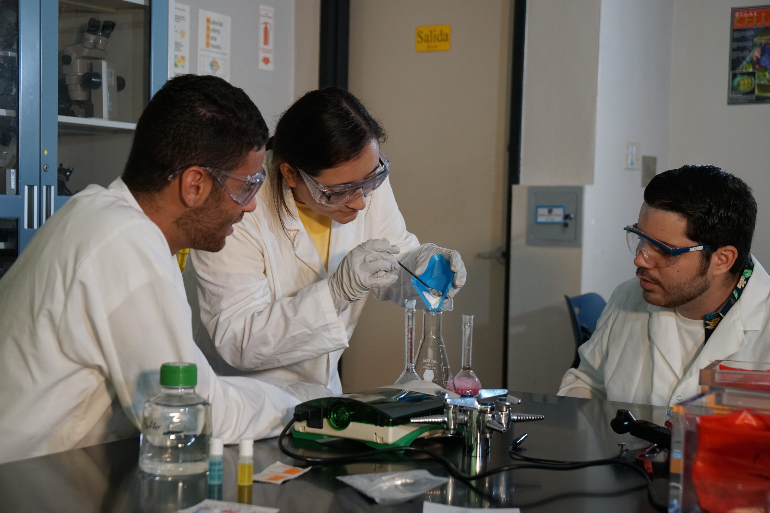 Tres estudiantes trabajando en grupo en un laboratorio. Uno está virtiendo un material pulverizado dentro de un matraz.