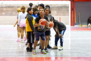 Jóvenes en fila en una cancha de baloncesto, estan esperando su turno para lanzar la bola al canasto.
