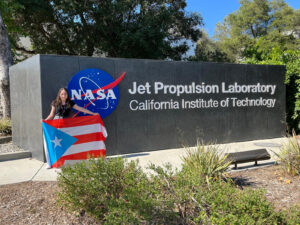 Arianna Rodríguez sosteniendo la bandera de Puerto Rico frente a un letrero de la NASA que lee "Jet Propulsion Laoratory"