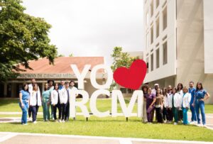 Grupo de estudiantes frente al edificio de Ciencias Médicas, en el centro está un letrero grande que lee "Yo Amo RCM"