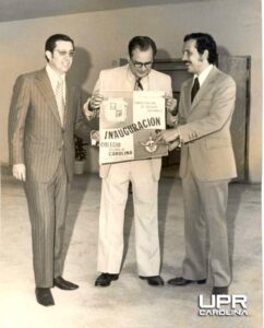 Foto en blanco y negro, 3 personas mostrando un afiche de la Inauguración de la UPR en Carolina