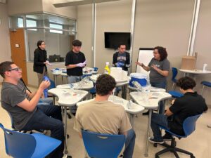 Varios estudiantes en un laboratorio con instrumentos en sus mesas. Profesora dando instrucciones.